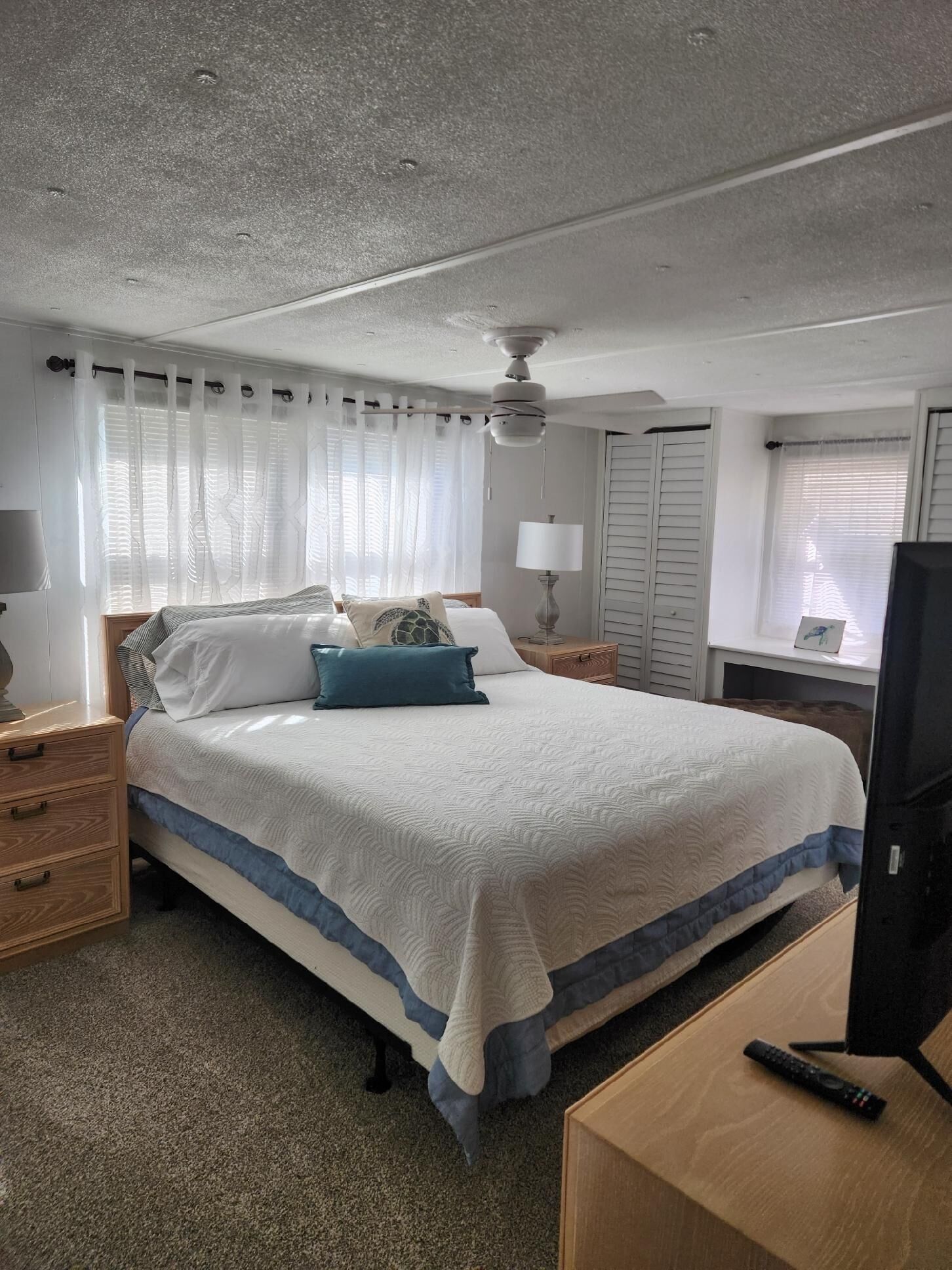 A hotel room with a bed, a ceiling fan, a nightstand, a lamp, a TV, and a window with curtains.