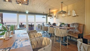 A modern dining area with a wooden ceiling, pendant lights, and a view of the sea through large windows. - Stunning home in Lembruch/Dümmer See with sauna (Lembruch/Dümmer See)