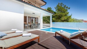 A poolside area with lounge chairs and a table, a covered patio, and a view of trees and a building in the background.