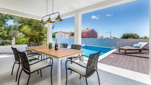 A covered outdoor area with a pool, wooden table, and chairs, overlooking a residential area.