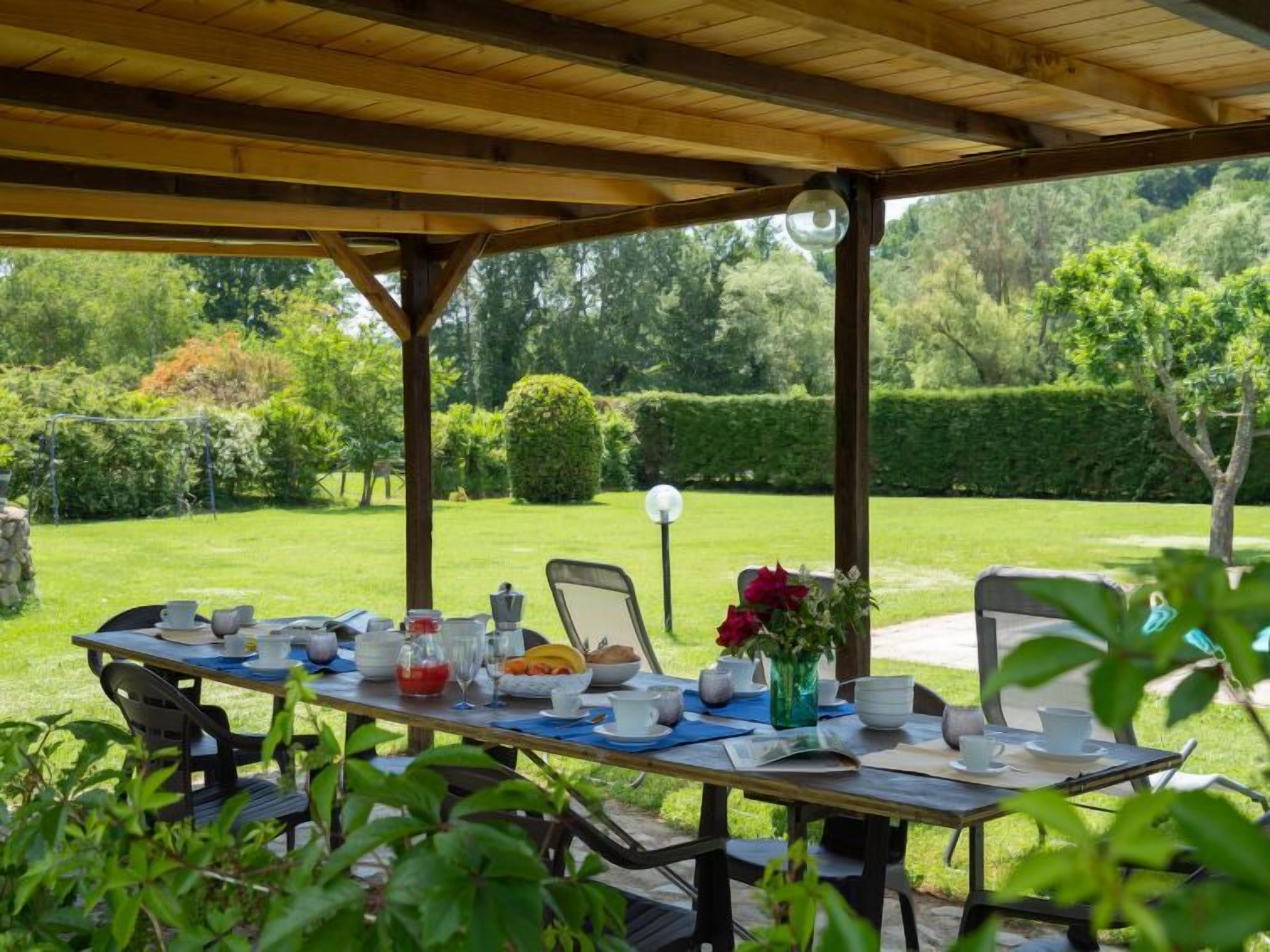 Un espacio de comedor al aire libre con una pérgola de madera, una mesa dispuesta con platos, tazas y un jarrón de flores.