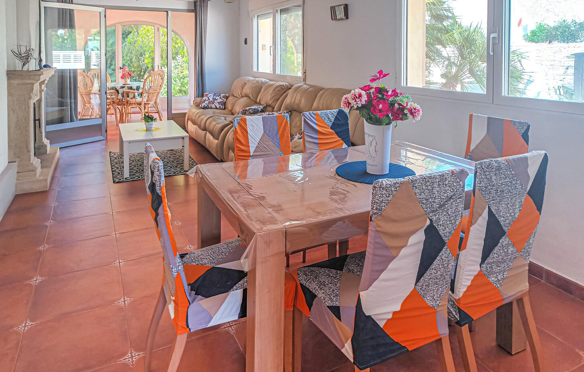 A dining area with a table set for four, surrounded by chairs with patterned cushions. A sofa and a window with a view of greenery are visible in the background.