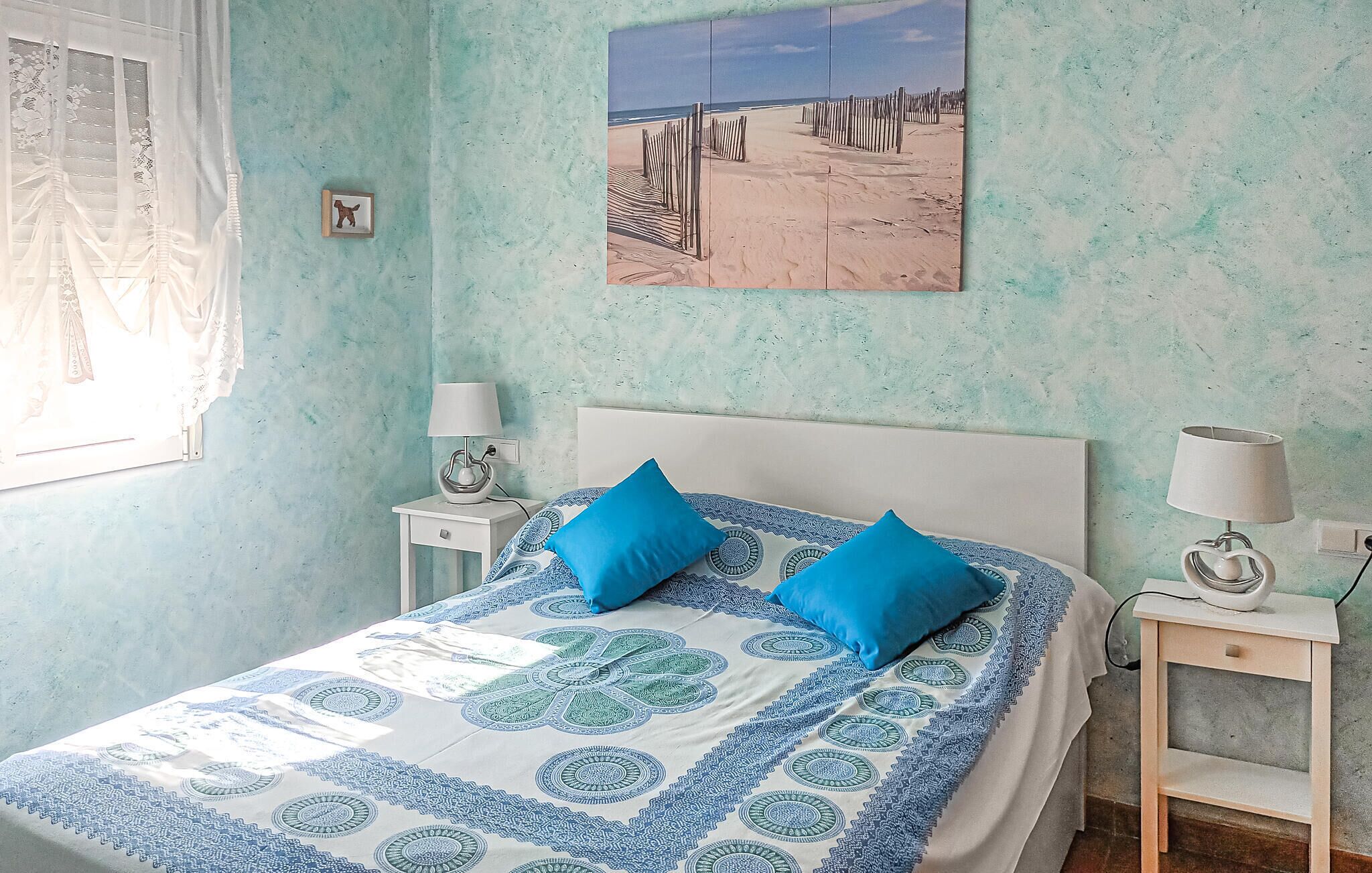 Un dormitorio con cama, dos mesitas de noche, una ventana con cortinas translúcidas, un cuadro con motivos playeros y una colcha estampada.