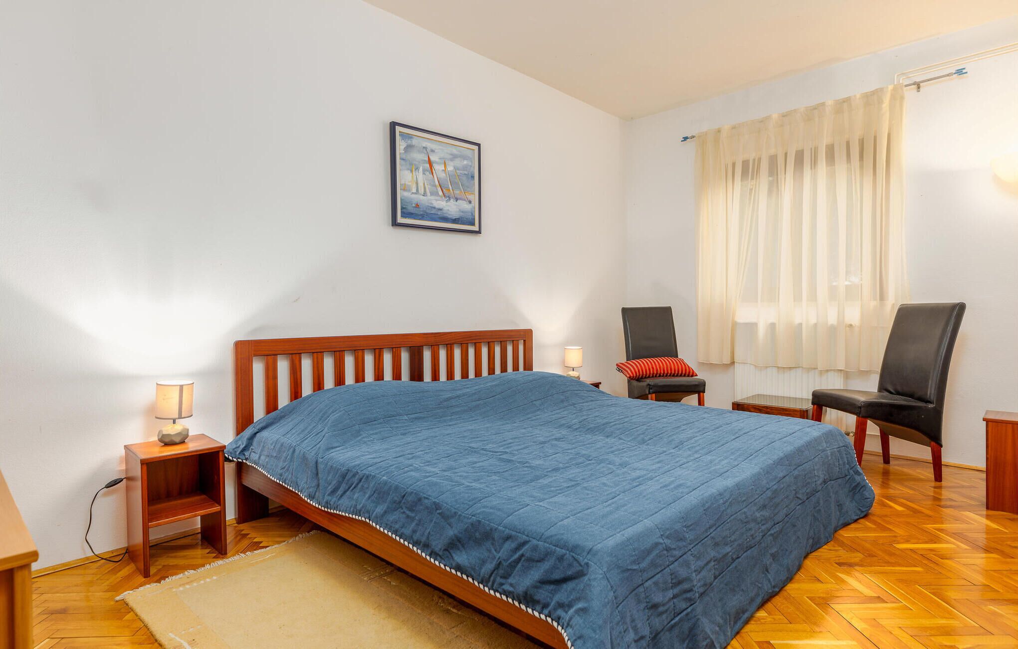 A bedroom with a wooden bed, a nightstand, a chair, a small table, a painting on the wall, and a window with curtains.