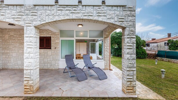 A covered outdoor seating area with two lounge chairs, a table, and a view of the house interior through a glass door.