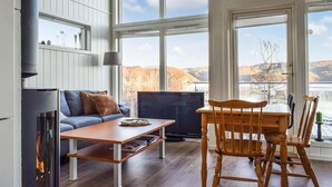 En hyggelig stue med pejs, sofa, træbord og fjernsyn. Store vinduer giver en smuk udsigt over bakker og et vandområde.