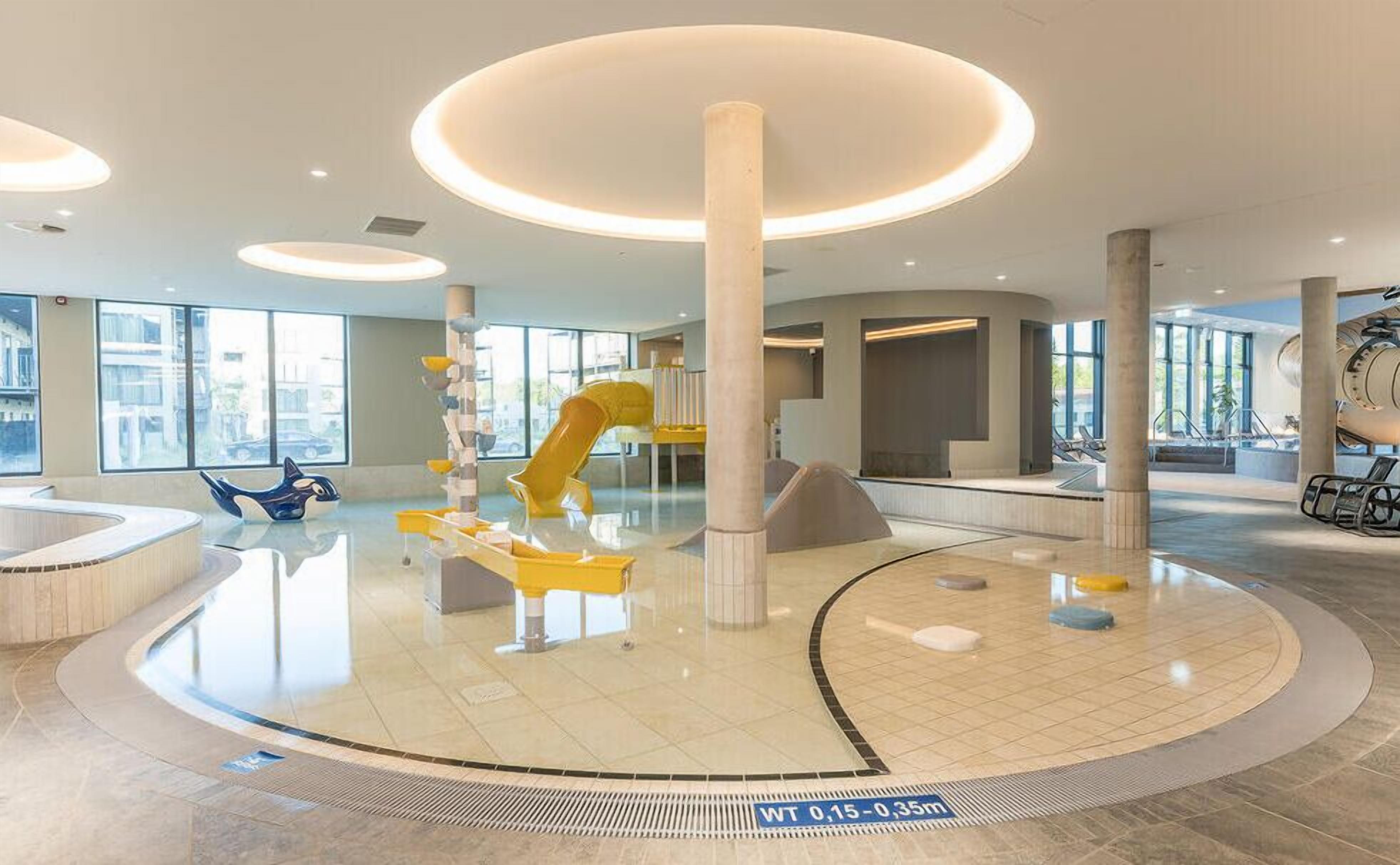 Un vaste espace intérieur doté d’un aménagement circulaire central, comprenant une aire de jeux avec des toboggans et un espace détente avec des chaises.