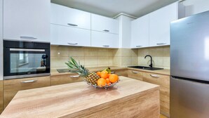 Eine moderne Küche mit weißen Schränken, einem Edelstahl-Kühlschrank, einem Einbauofen und einer Holzausziehtafel mit Obstschale.