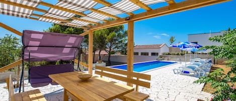 A wooden pergola with a pool, outdoor seating, and a clear blue sky.