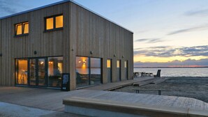 A modern wooden building with large windows, a wooden deck, and a view of the water at sunset. - Amazing home in Lembruch/Dümmer See with sauna (Lembruch/Dümmer See)