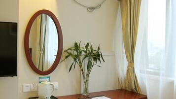 A hotel room with a desk, a mirror, a potted plant, and a TV.