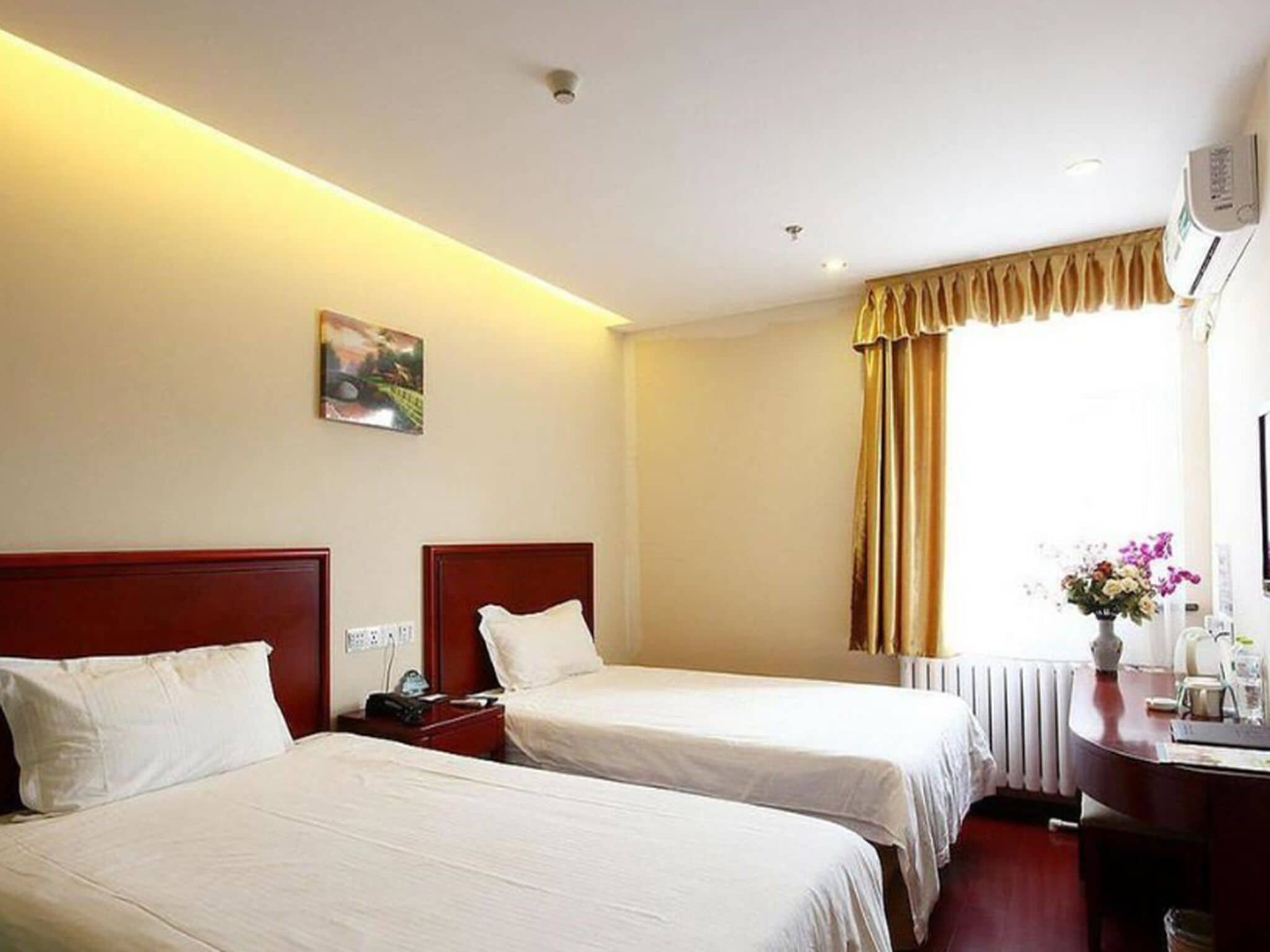 Una camera d'albergo con due letti singoli, un comodino con una lampada, un quadro appeso al muro, una finestra con tende e un termosifone.