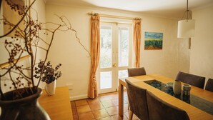 A dining area with a wooden table, chairs, a vase with dried flowers, a painting on the wall, and a window with curtains.