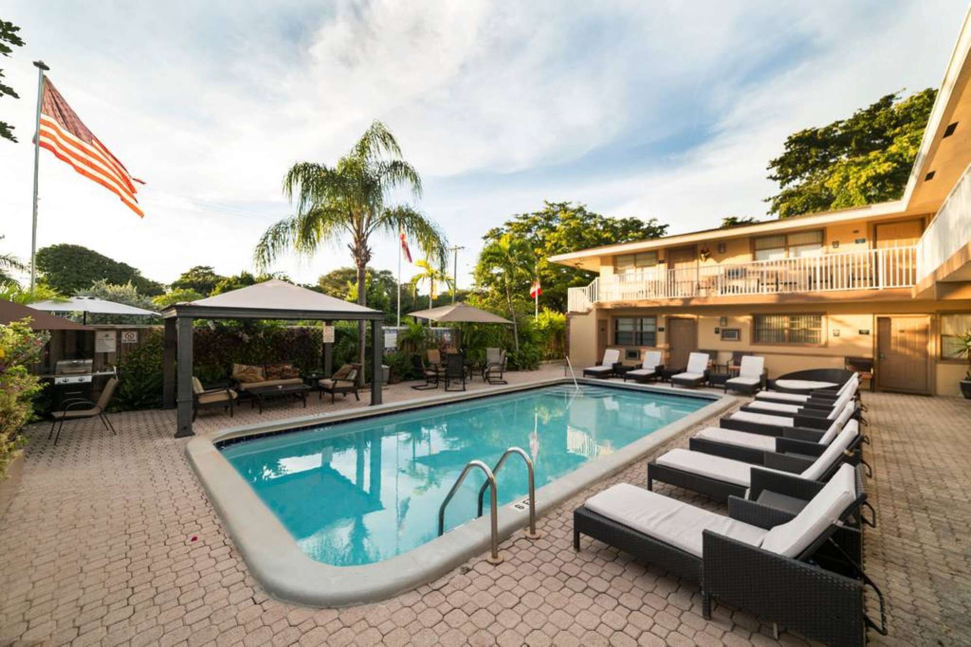 Ein Pool mit Liegestühlen und einer überdachten Sitzecke, umgeben von Grünflächen und einem Gebäude mit Balkon.