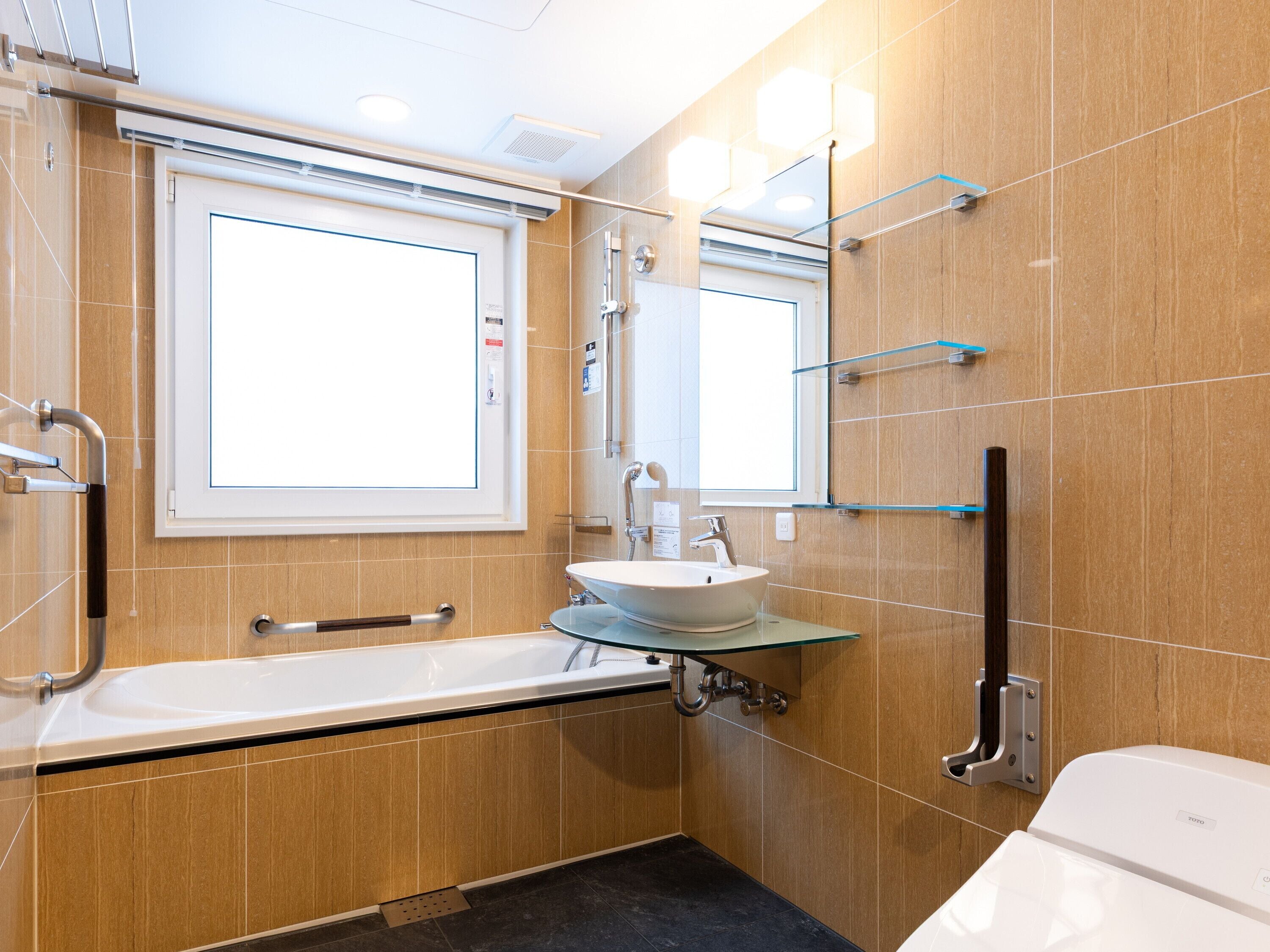 A modern bathroom with a bathtub, a sink, and a toilet. There are grab bars and a window above the sink.