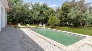 Una piscina rectangular con borde de piedras, rodeada de césped y árboles. Hay cuatro sillas de jardín blancas alineadas a lo largo de la piscina.