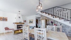 A spacious living room with a fireplace, wooden dining table, and chairs. A staircase with a wrought iron railing leads to an upper level. - Holiday Home Rustic (Muc)