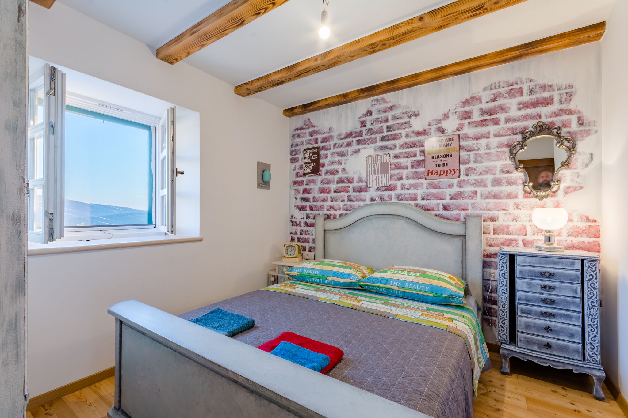 A bedroom with a bed, a window, a brick wall, a mirror, and a dresser.