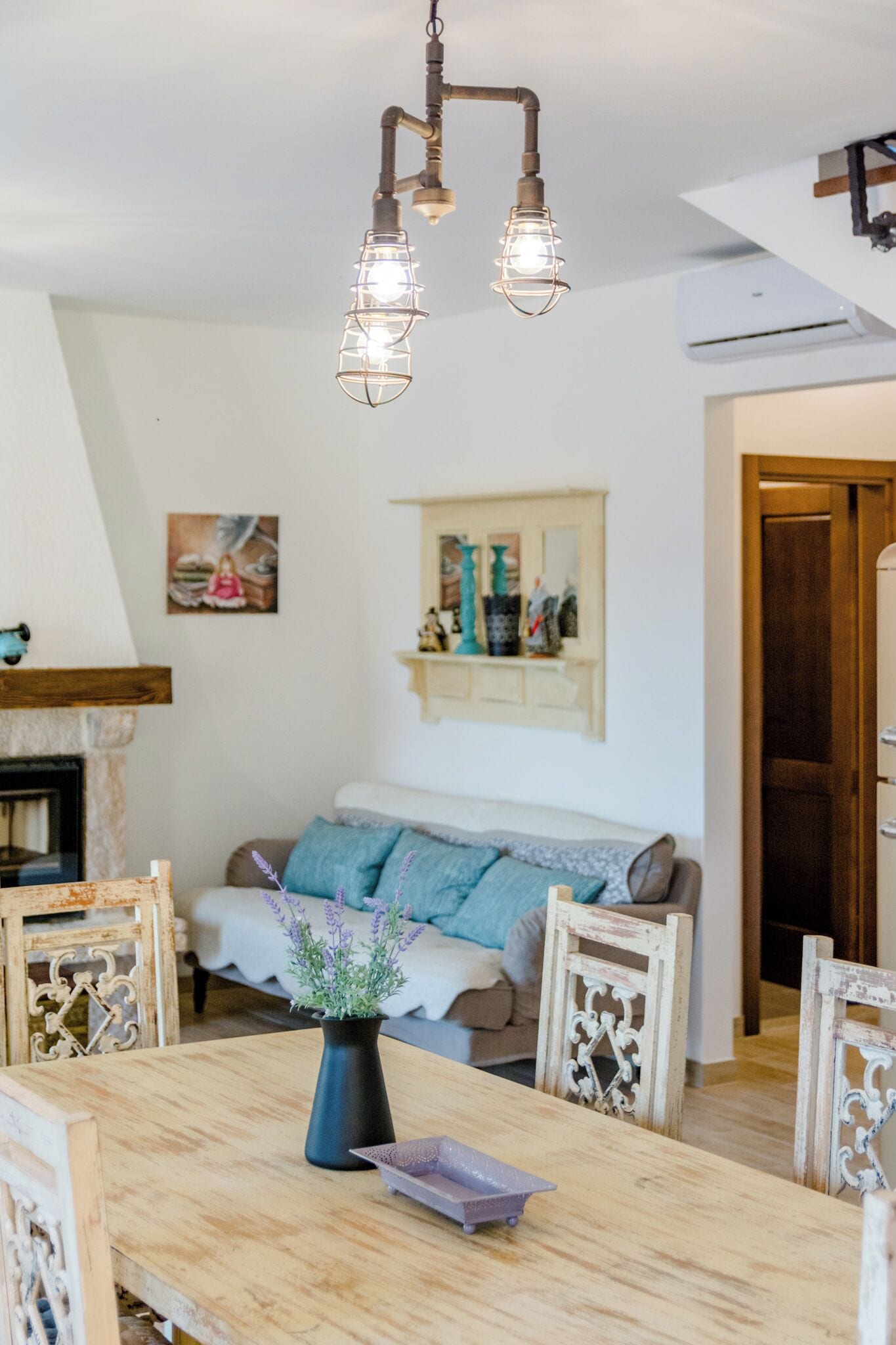 A dining area with a wooden table, chairs, and a vase of flowers. There is a fireplace and a sofa in the background.