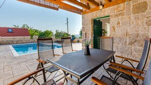 A covered outdoor seating area with a table and chairs, a stone wall, and a view of a swimming pool. - Holiday Home Rustic (Muc)