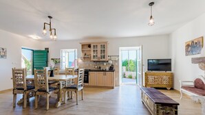 A spacious living area with a wooden dining table and chairs, a kitchen with wooden cabinets, a television, and a bench. - Holiday Home Rustic (Muc)