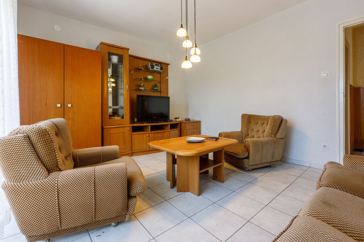 Ein Wohnzimmer mit Sofa, Sessel und einem kleinen Holztisch. Ein Fernseher und eine Schrank mit Regalen sind ebenfalls vorhanden.