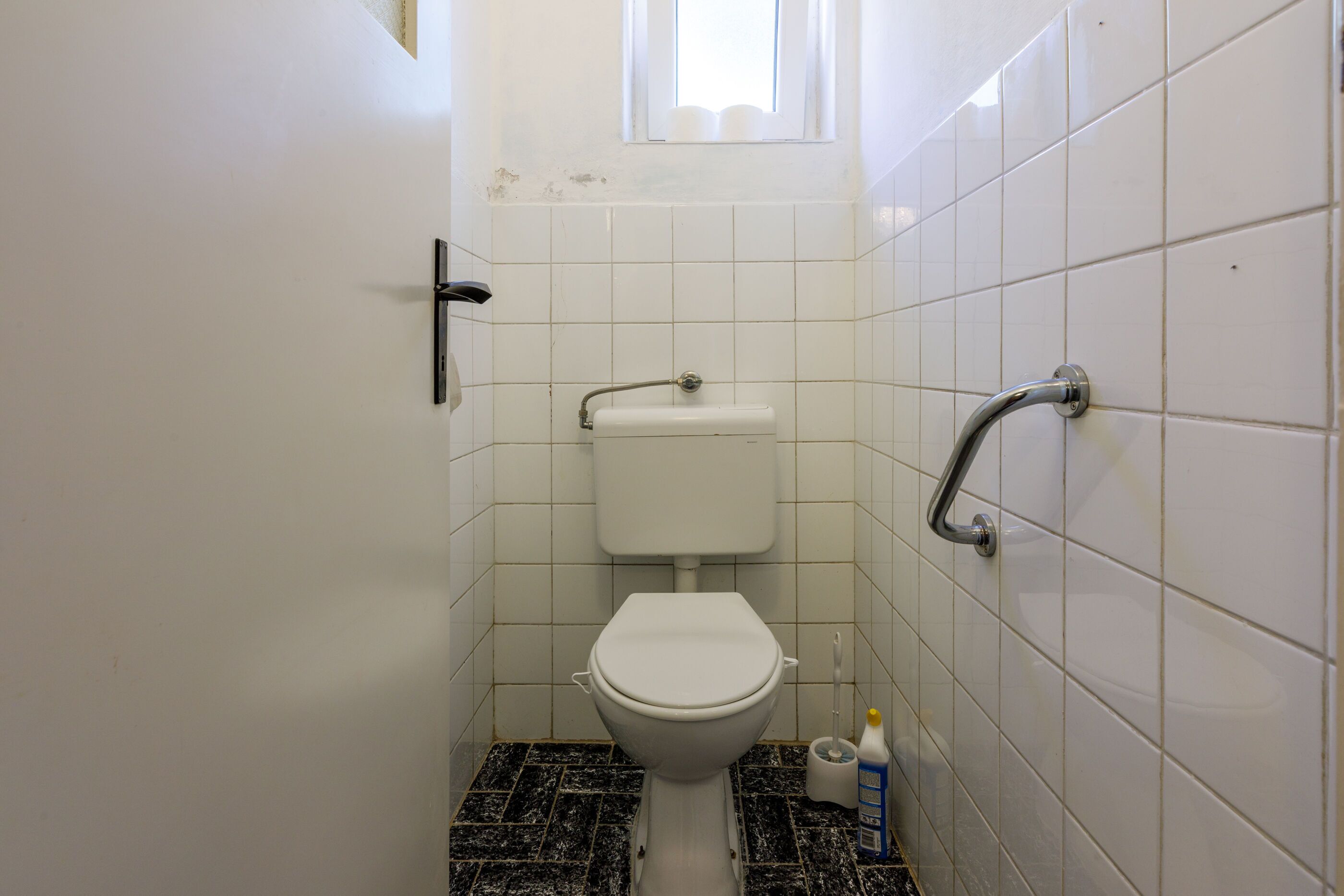 A small bathroom with a toilet, a handrail, and a window.