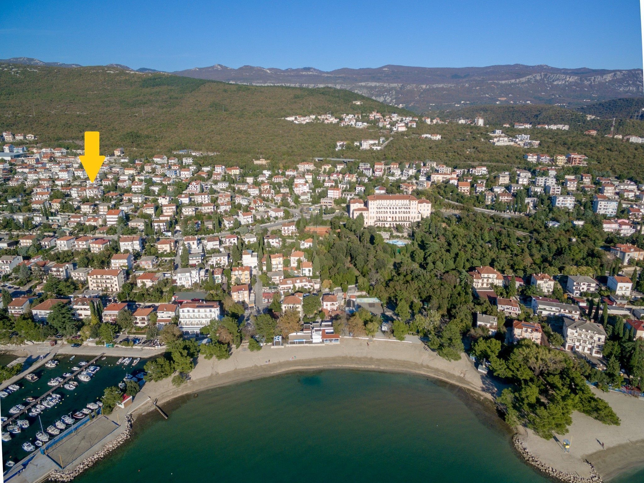 Vista aérea de un pueblo costero con edificios residenciales, un puerto deportivo y un edificio grande en una colina.