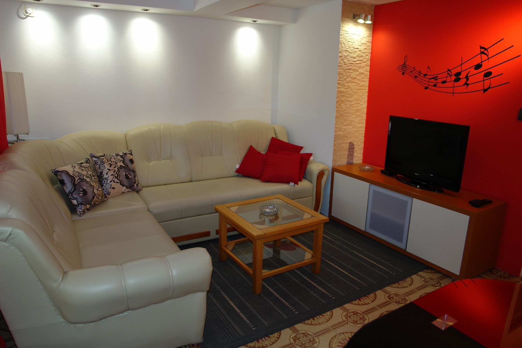 Una sala de estar con un sofá beige, cojines rojos, una mesa de centro de cristal, un televisor de pantalla plana y una pared de acento roja con un diseño de nota musical.