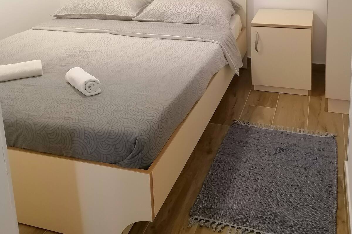 Un letto rifatto con cura, con una coperta grigia a motivi e cuscini, un comodino con una lampada e un tappeto grigio intrecciato sul pavimento in legno.