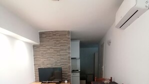 Una habitación funcional con una cama, un televisor sobre un soporte de madera, una mesa de comedor con sillas y una kitchenette al fondo.