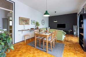 A modern living room with a wooden dining table, green sofa, and a TV mounted on the wall. - Apartment Suzy (Crikvenica)