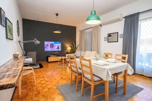 A modern living room with a dining area, wooden furniture, a television, and a large window with curtains.