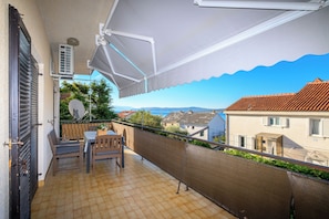 A balcony with a table and chairs, a view of the sea, and a building with a tiled roof. - Apartment Suzy (Dramalj)