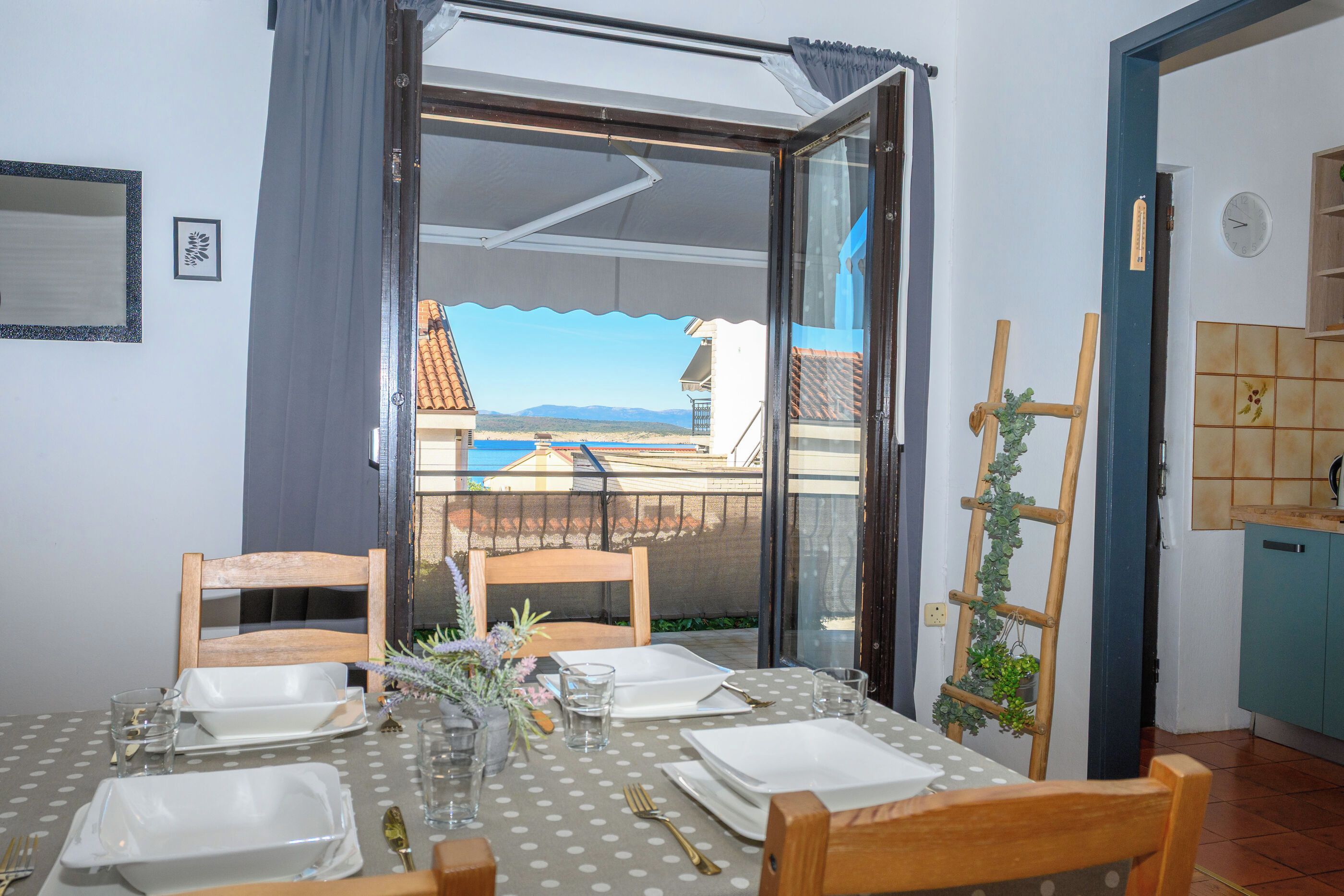 A dining area with a table set for four, a view of the sea and buildings, and a kitchen area with a wooden ladder and tiles.