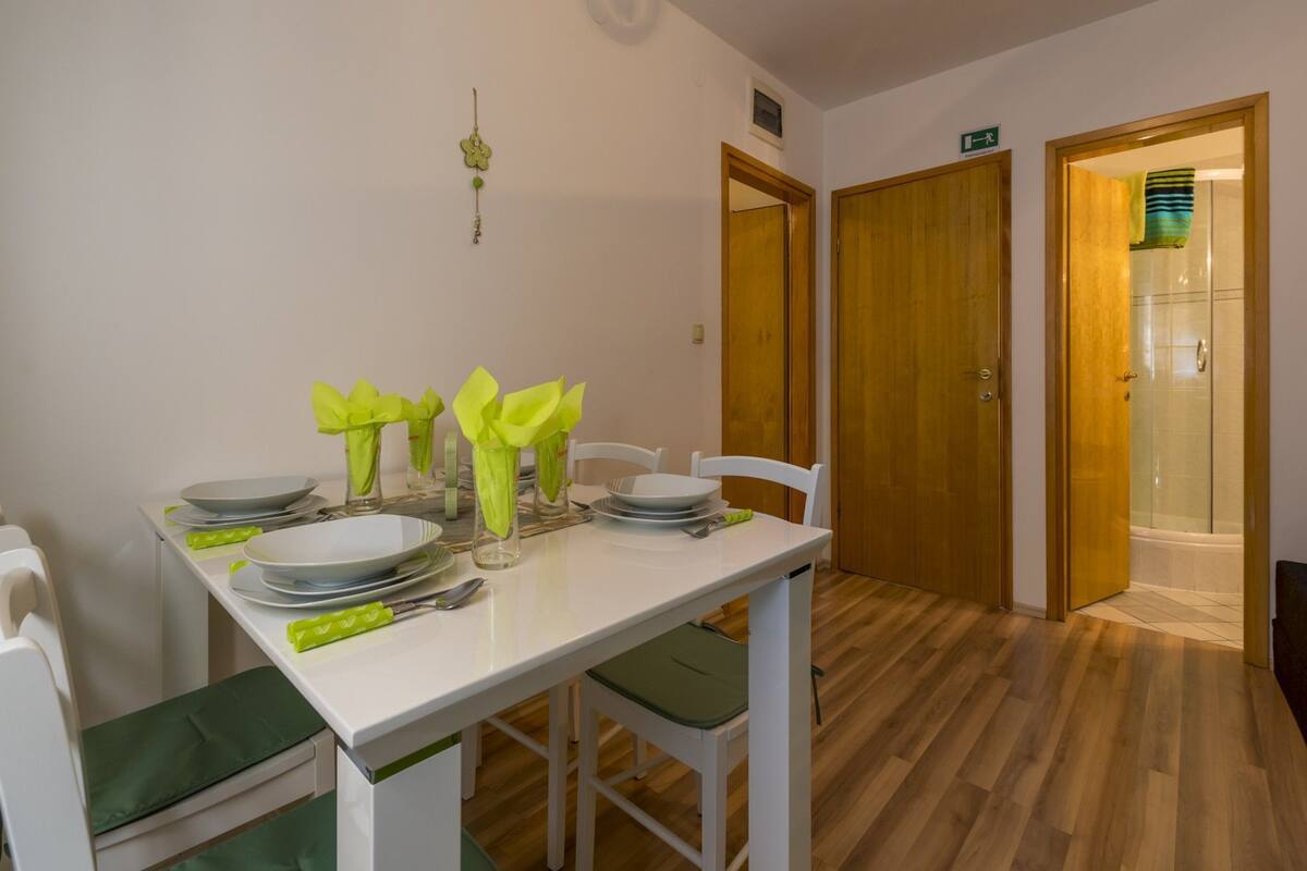 Un coin repas avec une table blanche dressée pour quatre personnes, des coussins verts et un plancher en bois.