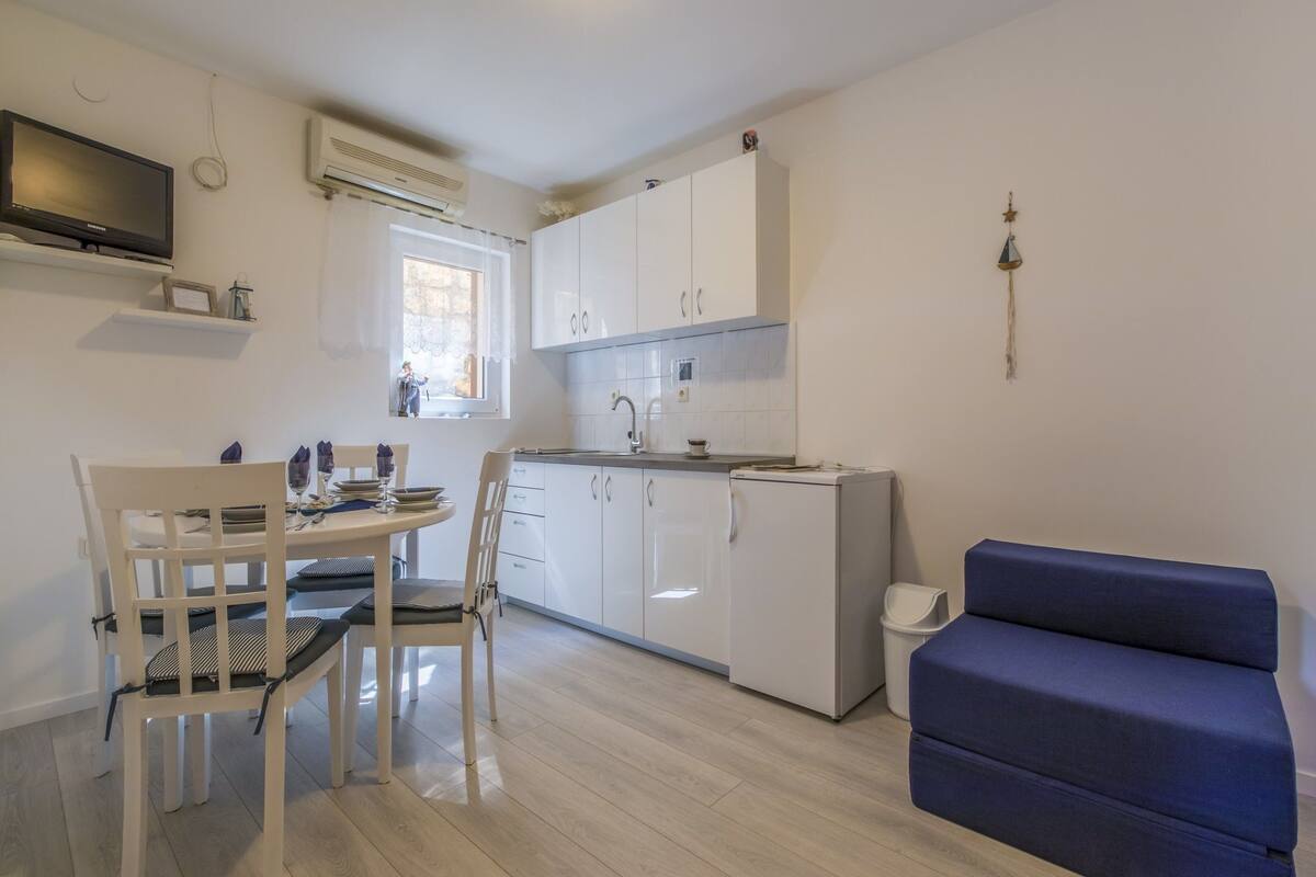 Une cuisine compacte avec des meubles blancs, un évier intégré et un petit réfrigérateur. On y trouve une table à manger avec des chaises, ainsi qu’un canapé bleu dans l’espace salon.