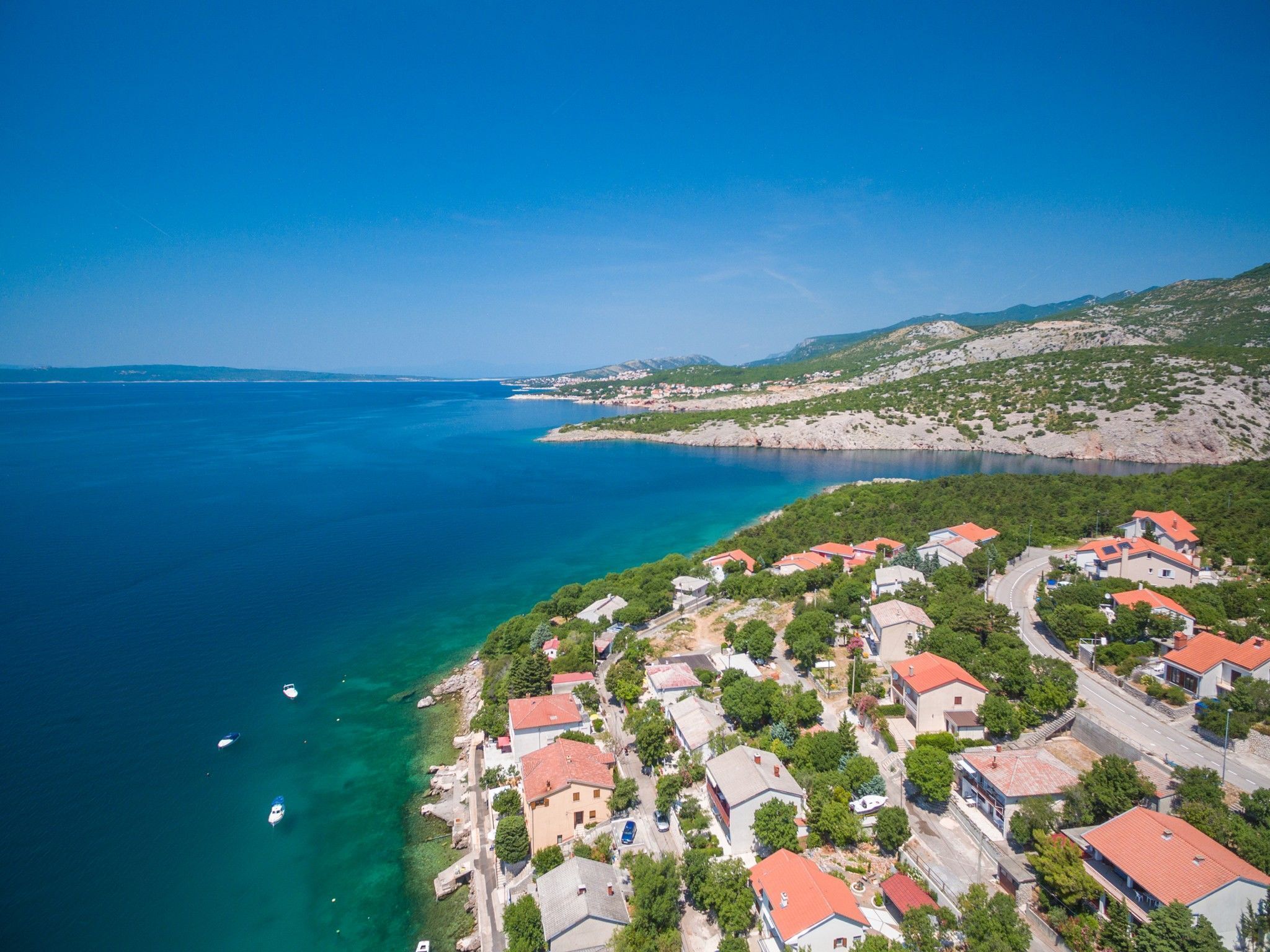 An aerial view of a coastal village with houses along the shoreline, a clear blue sea, and a hilly landscape in the background.