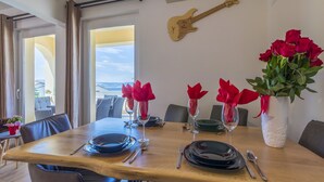 Ein Esstisch ist gedeckt mit Tellern, Besteck und roten Blumen in Vasen. An der Wand hängt eine Gitarre und durch das Fenster ist eine Aussicht zu sehen.