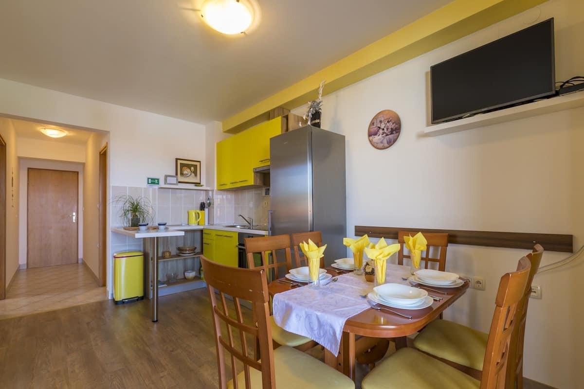 Un coin repas avec une table en bois dressée pour quatre personnes, des chaises jaunes et une cuisine en arrière-plan.