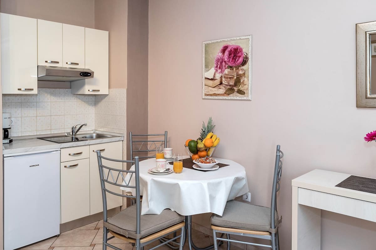A compact kitchen with white cabinets, a stainless steel sink, and a small refrigerator. A dining table with a white tablecloth and chairs is set for breakfast.