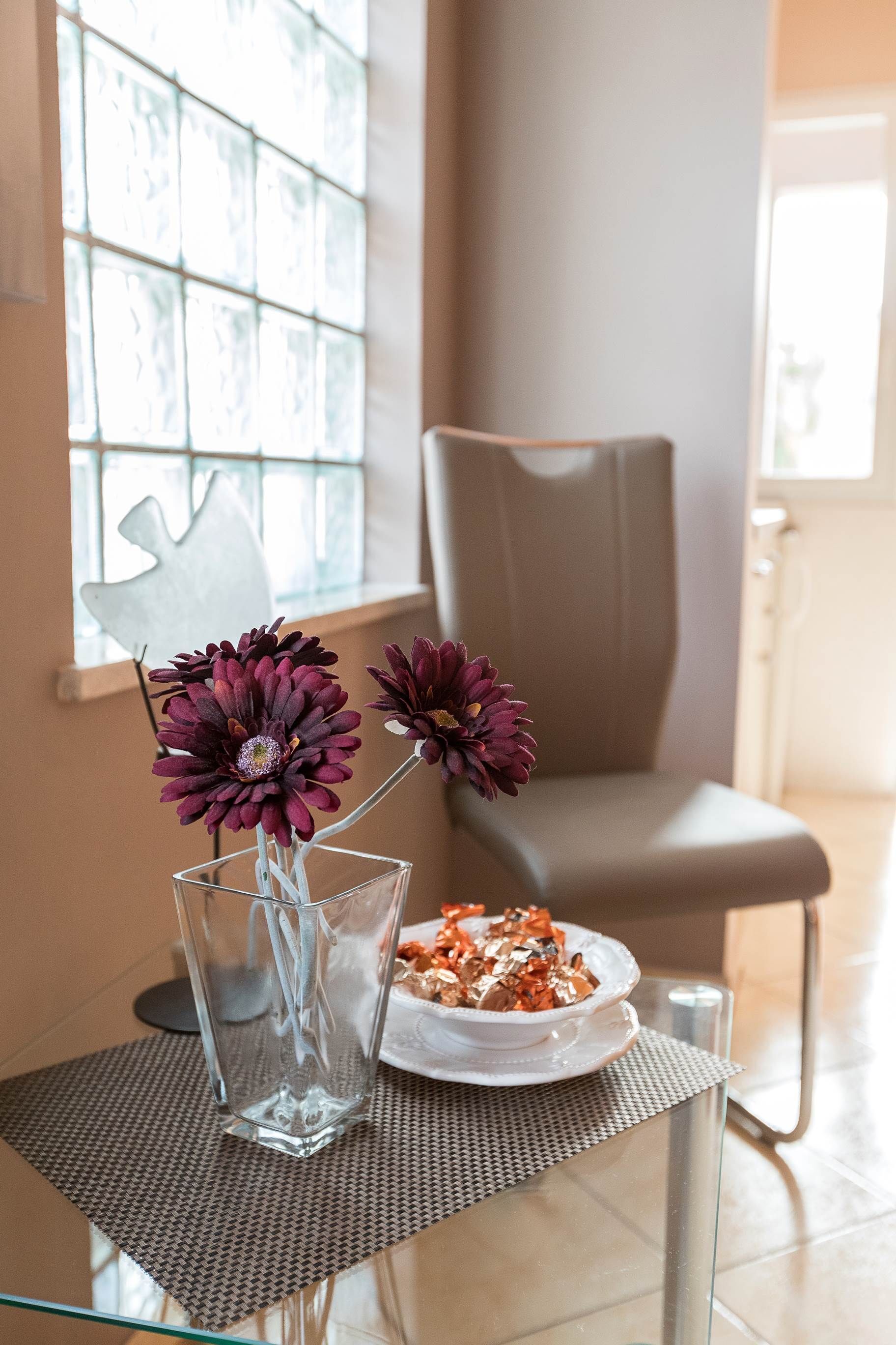 A glass table with a vase of purple flowers and a plate of food, a chair, and a window with a decorative element.