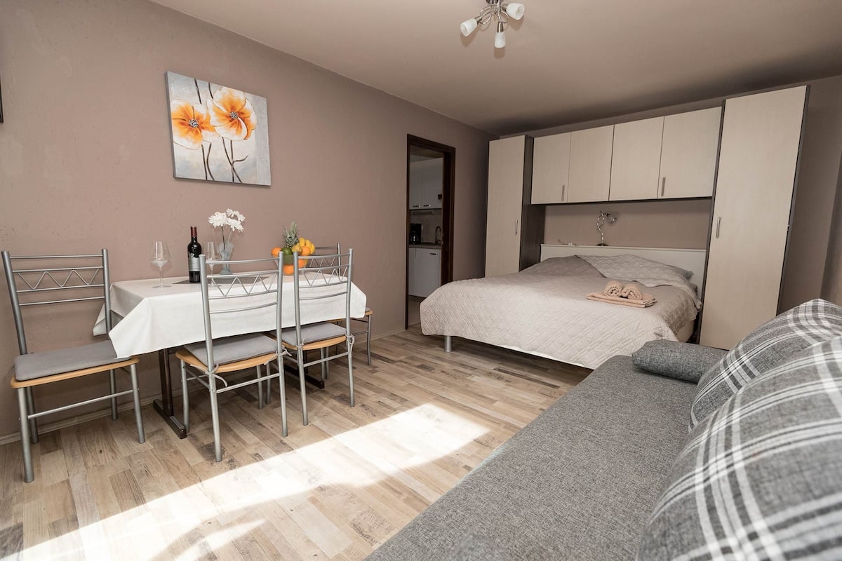 A bedroom with a bed, a sofa, a dining table with chairs, and a kitchen area in the background.