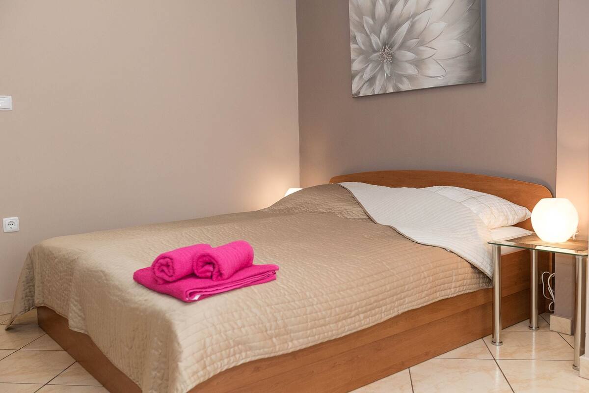 A neatly made bed with a beige quilt, a pink towel, and a bedside lamp.