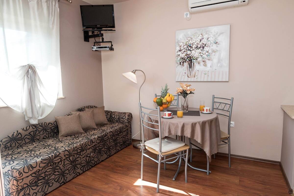 A cozy living area with a sofa, a dining table with chairs, a TV mounted on the wall, and a painting above the table.