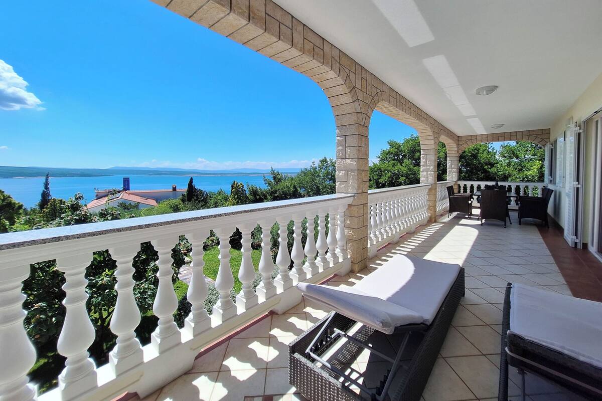A covered balcony with a view of a lake and distant mountains, featuring a table and chairs, and a covered grill area.