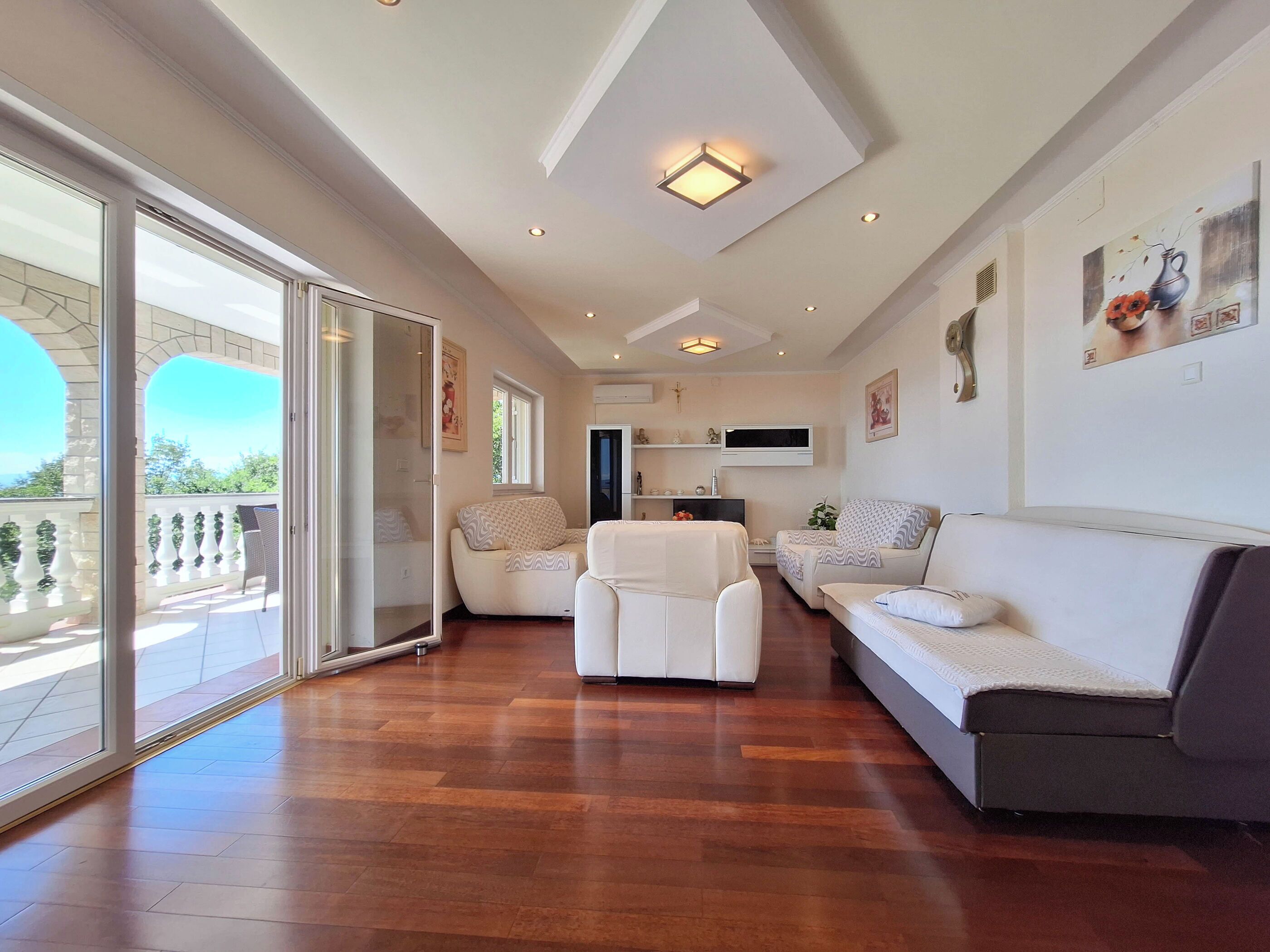 A spacious living area with a large sliding glass door leading to a balcony, a flat-screen TV mounted on the wall, and a ceiling with recessed lighting.