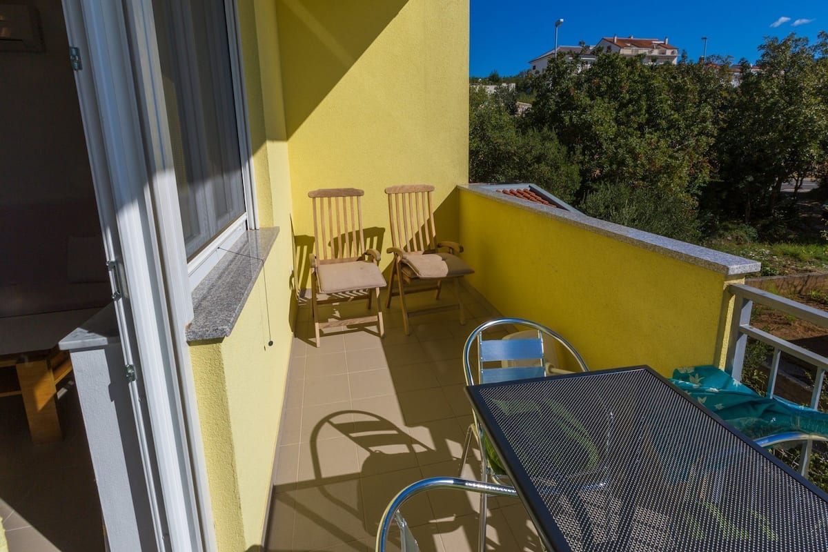 A balcony with yellow walls, two wooden chairs, a metal table, and a view of greenery and buildings in the background.