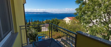 A balcony with a metal table and chairs, overlooking a coastal view with a clear sky.