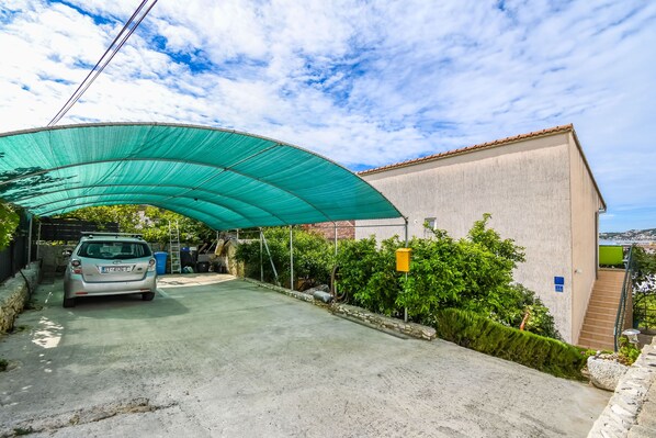 A car under a green canopy, a building with a stone wall, and a clear sky. - Apartments Salov (Trogir)